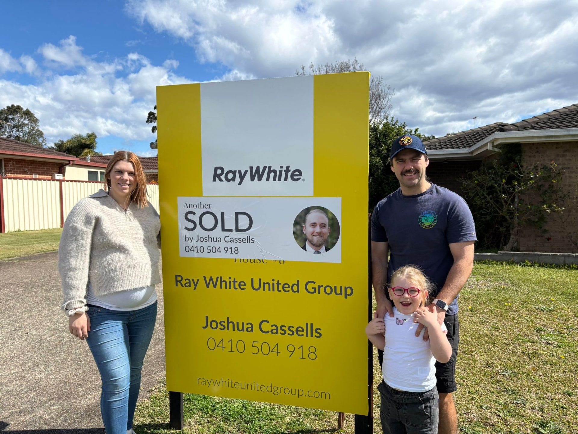 A family with their daughter beside a Sold sign at their home