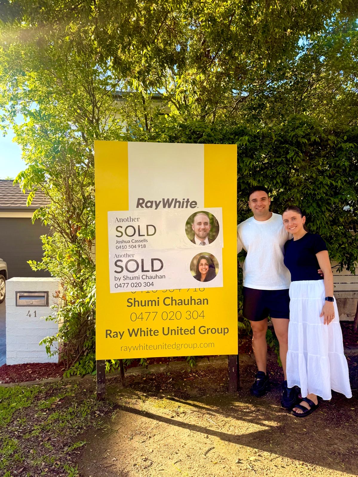 Two sellers beside a Sold sign at their home
